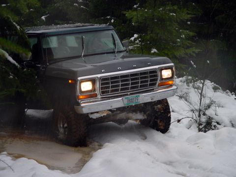 1979 Ford Bronco - A dark green 1979 Ford Bronco navigates a snowy, wooded terrain, its headlights illuminating the path ahead.