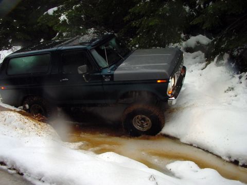 1979 Ford Bronco - A dark green Ford Bronco navigates a snowy, muddy trail, splashing water as it goes.