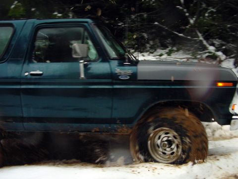 1979 Ford Bronco - A teal Ford Bronco navigates a snowy and muddy terrain, showing its side profile.
