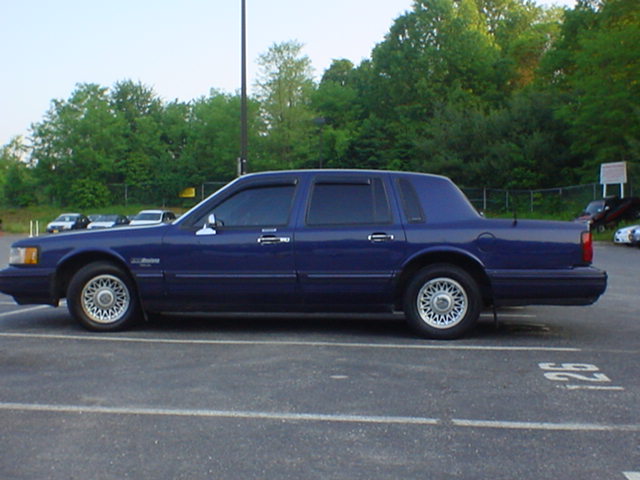Side profile of dark blue Lincoln Town Car sedan on chrome lace alloy wheels in a parking lot