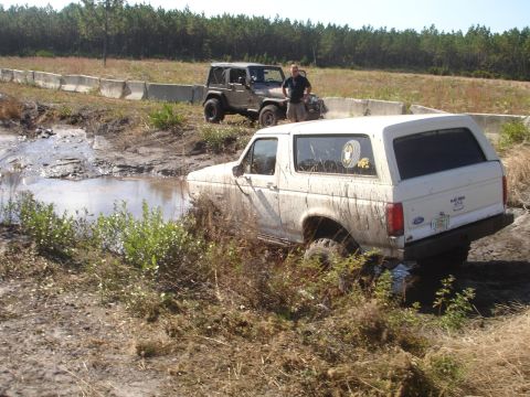 1989 Ford Bronco - A white 1989 Ford Bronco navigates a muddy obstacle course, with another off-road vehicle visible in the background.