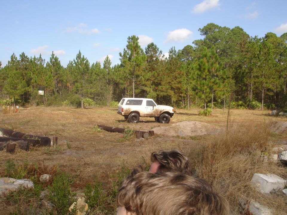 White SUV parked in a grassy, wooded area with some dirt mounds. Person in foreground