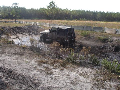 1989 Ford Bronco - A Jeep Wrangler struggles through a muddy obstacle course, kicking up dirt and water.