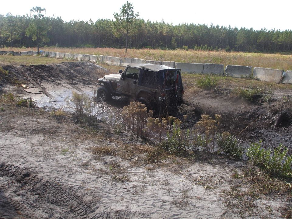 Mud-splattered Jeep Wrangler navigating a muddy track with trees and concrete barriers in the background