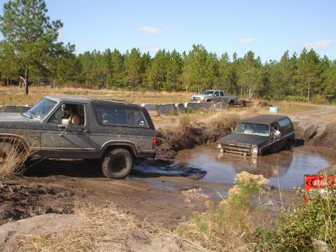 1989 Ford Bronco - Two SUVs navigate a muddy off-road course on a sunny day.