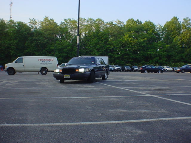 Dark blue Lincoln Town Car with headlights on in an empty parking lot beside a Freehold Nissan van