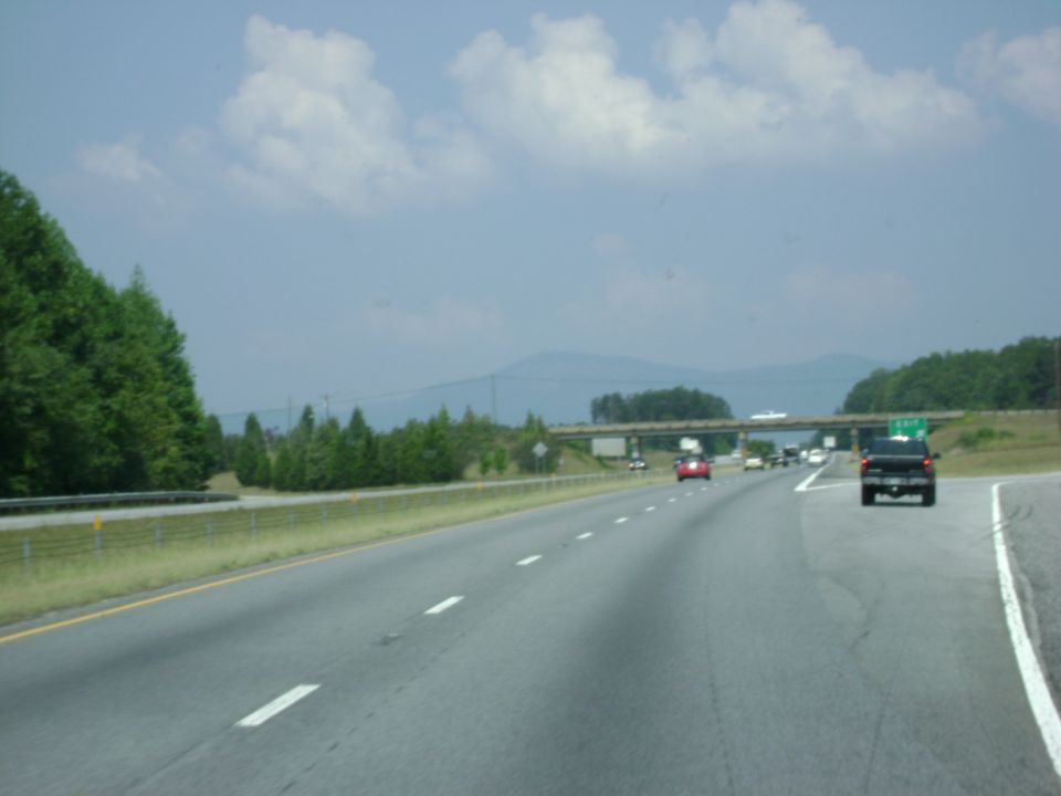 Interstate highway view with dark pickup and red car ahead, mountains and overpass in distance