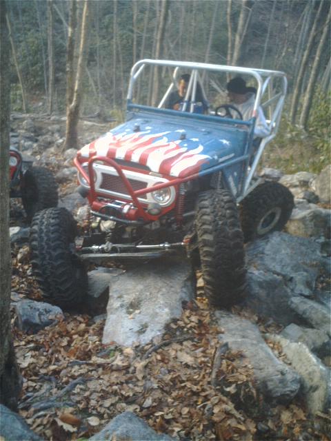 1992 Ford Mustang - An off-road vehicle with patriotic paint navigates a challenging rocky path in a wooded area.