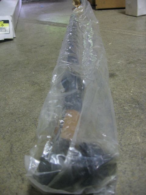 1989 Ford Bronco - A long black driveshaft still sealed in a clear plastic bag, laid lengthwise on a bare concrete shop floor with a cardboard box and shelvin…