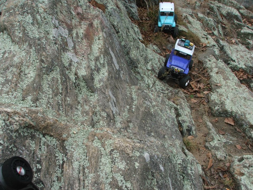 Two remote-control trucks, one blue and one light blue, climb a rocky, lichen-covered slope