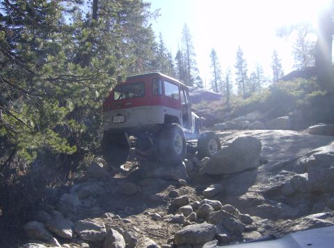 1986 Ford Bronco - Off-road vehicle navigating a difficult rocky path with trees and clear sky in the background.