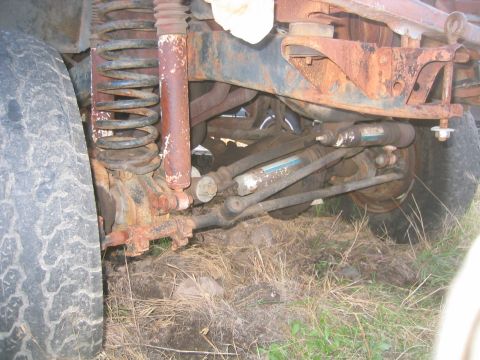 1973 Ford F-100 - Low side view under a truck's coil-sprung front suspension showing a surface-rusted frame rail, radius arm, dual shocks with blue bodies, t…