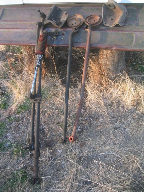 1973 Ford F-100 - Rusty coil-spring front suspension parts leaned against a weathered truck bed side outdoors: two radius arms, a track bar/steering link wit…