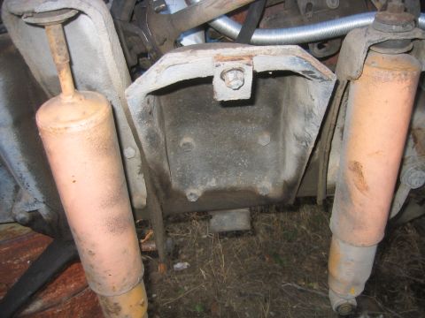 1973 Ford F-100 - Two dusty yellow shock absorbers with exposed chrome shafts and eyelet/stud ends stand propped beside a rusty stamped-steel coil-spring tow…