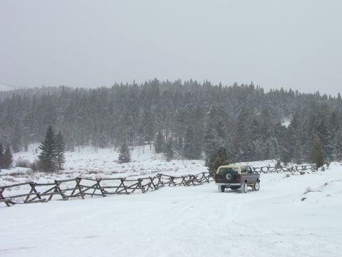 1979 Ford Bronco - A 1979 Ford Bronco drives along a snow-covered road bordered by a rustic wooden fence and evergreen trees.