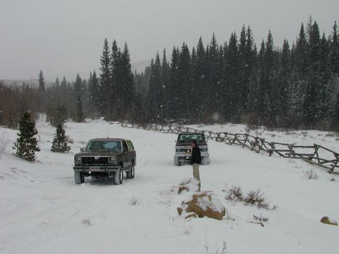 1979 Ford Bronco - Two SUVs navigate a snowy landscape, with a wooden fence and dense pine forest in the background.