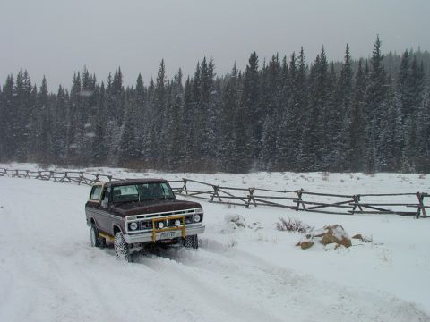 1979 Ford Bronco - A 1978 Ford Bronco navigates a snowy path, bordered by a rustic wooden fence and dense pine forest.