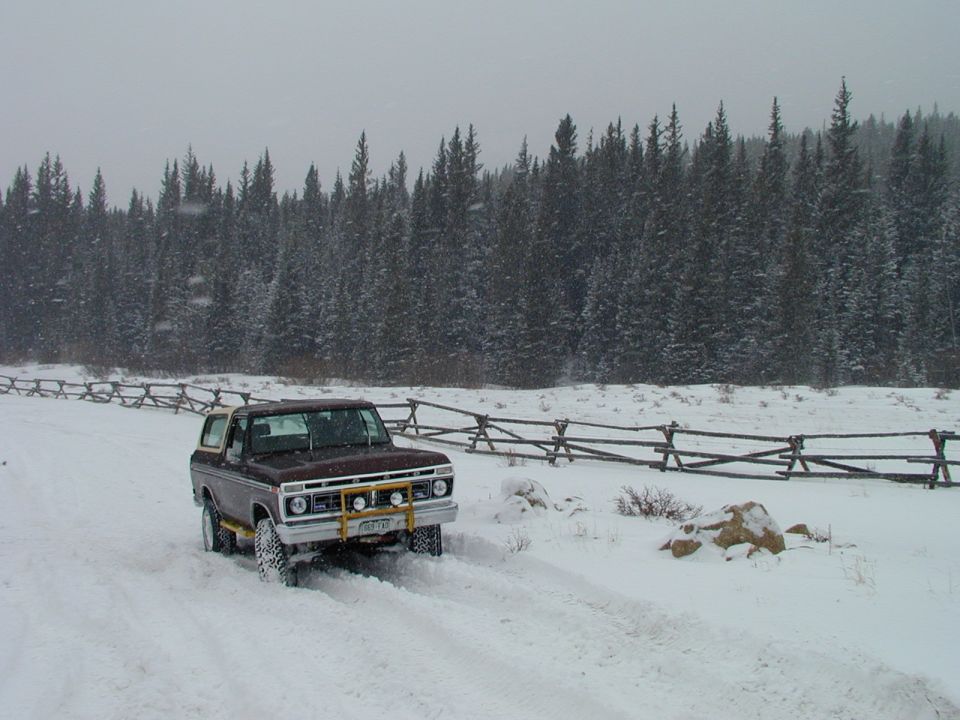 Brown 1978 Ford Bronco driving on a snowy road with a wooden fence and pine trees in the background