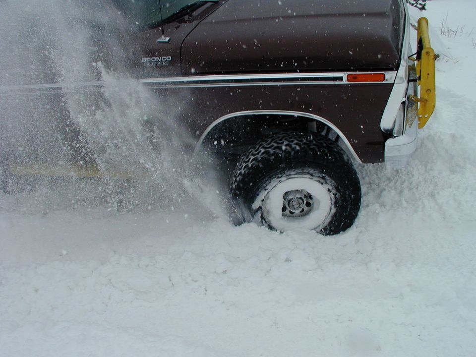 Brown 1978 Ford Bronco with yellow brush guard driving in deep snow, kicking up spray