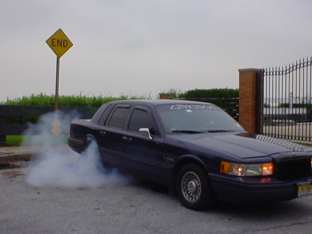 Dark blue Lincoln Town Car sedan doing a smoky burnout beside a yellow END sign