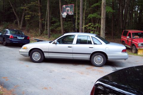 2001 Ford Crown Victoria - A silver 2001 Ford Crown Victoria is parked outdoors next to other vehicles and a basketball hoop.