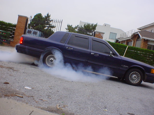 Dark blue sedan with tinted windows doing a burnout, emitting smoke from rear tires. Visible side profile and rear quarter