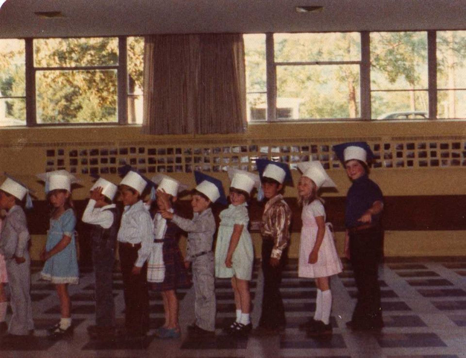 Children in graduation caps stand in a line indoors, with windows showing trees outside