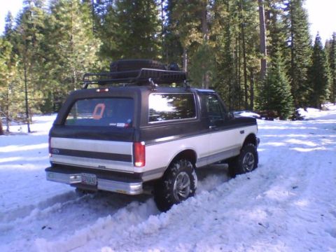 1993 Ford Bronco - A two-tone Ford Bronco drives through snow-covered terrain with a roof rack and spare tire.