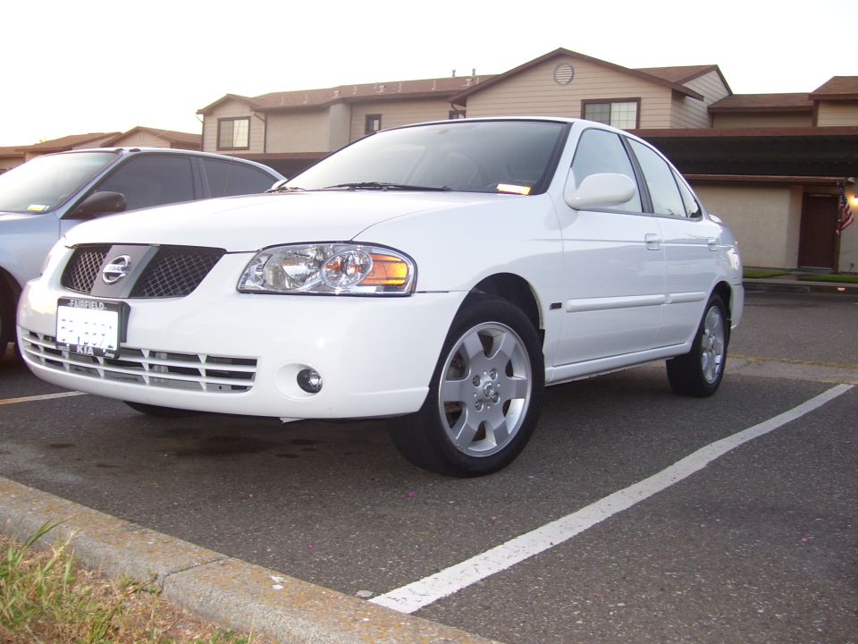White Nissan Sentra parked in a lot, another car partially visible to the left