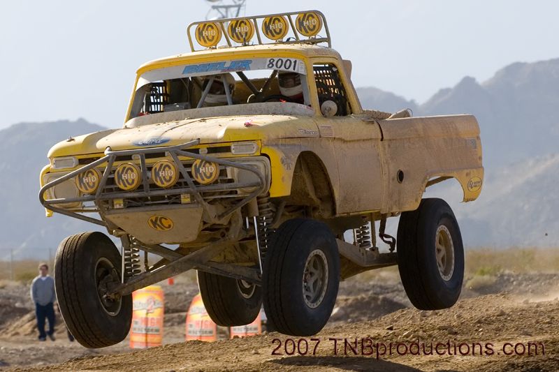 Yellow off-road truck mid-air, number 8001 on windshield banner. KC lights on bumper and roof