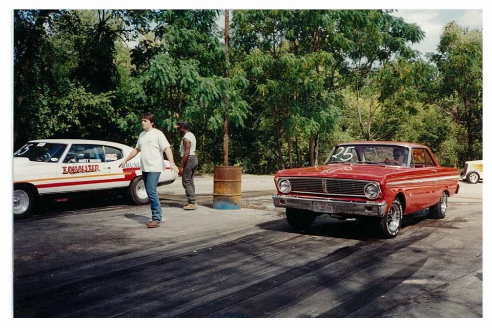 Red 1965 Ford Falcon coupe with '25' on windshield at a drag strip, white race car and people nearby