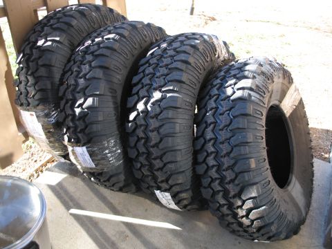 1995 Ford Bronco - Four new mud-terrain off-road tires with aggressive lug tread stand in a row on a concrete surface, some still wrapped in plastic with pape…