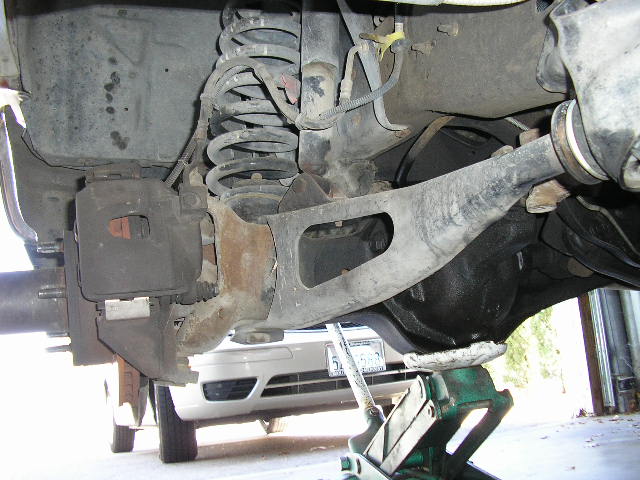 Rear suspension components including coil spring, brake caliper, differential housing, and control arm. A floor jack supports the differential