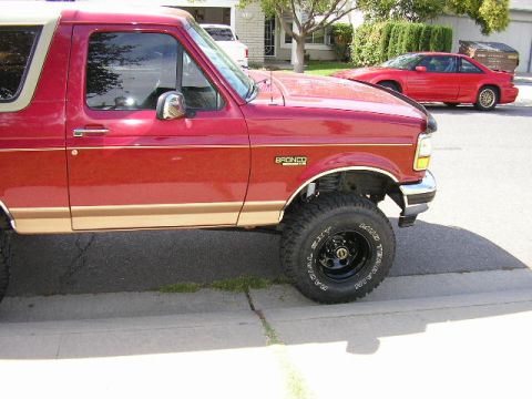 1994 Ford Bronco - Side view of the front half of a red and tan two-tone Ford Bronco parked at a residential curb, showing the "Bronco" fender badge, tan lowe…