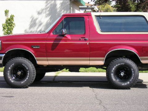 1994 Ford Bronco - A two-tone red and tan Ford Bronco full-size SUV shown in side profile, lifted with black steel wheels and white-lettered all-terrain tires…