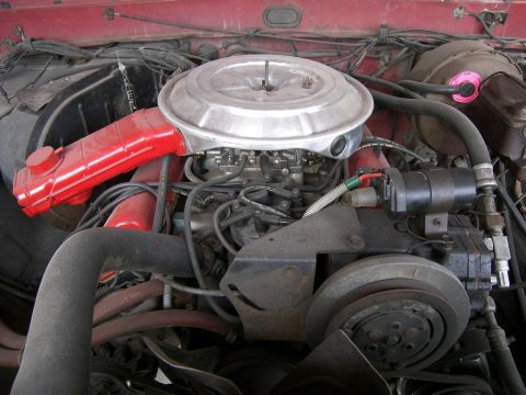 1978 Ford Bronco - Engine compartment of a red vehicle showing a carbureted V8 with chrome air cleaner, red spark-plug wires and accessory pulleys.