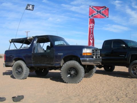 1994 Ford Bronco - A lifted 1994 Ford Bronco and a black Toyota Tacoma are parked on a sandy terrain under a blue sky with flags flying.