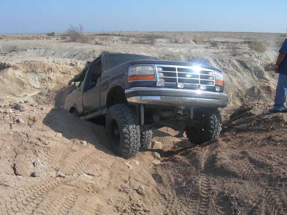 Lifted 1990s Ford Bronco climbing out of a rocky desert wash, front three-quarter view