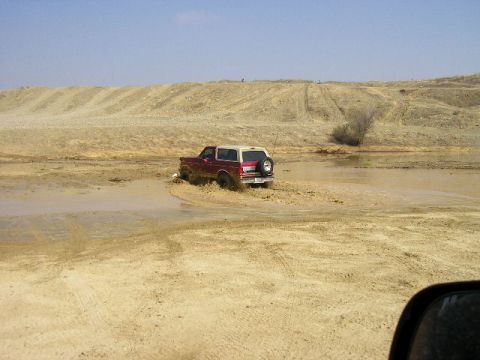 1994 Ford Bronco - A red and white SUV navigates through a muddy terrain under a clear blue sky.