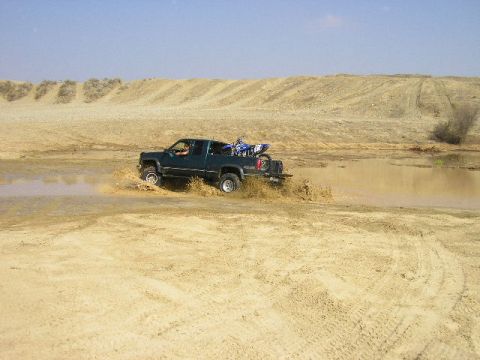 1994 Ford Bronco - A green pickup truck with a dirt bike in its bed splashes through a muddy puddle in a dry, sandy landscape.