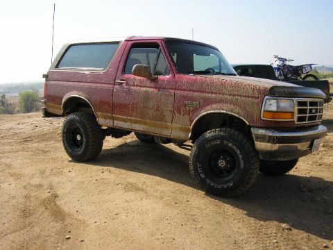 1994 Ford Bronco - This muddy 1994 Ford Bronco is seen off-road, with a dirt bike and another vehicle in the background.