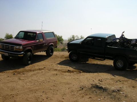 1994 Ford Bronco - Two trucks are parked on a dirt lot under a clear sky. The Bronco is maroon and the pickup is black.