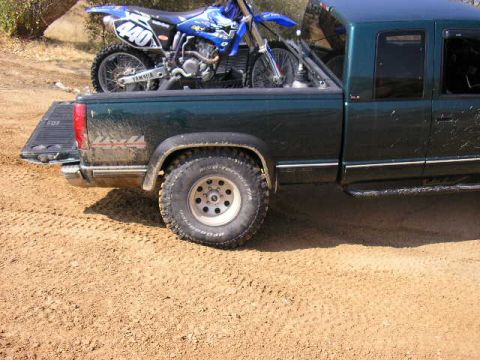 1994 Ford Bronco - A dark green pickup truck carries a blue dirt bike in its bed, parked on a dirt surface.