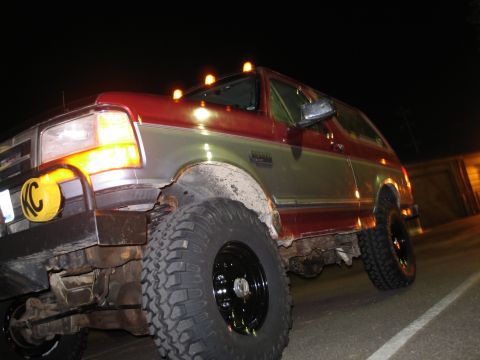 1995 Ford Bronco - Low-angle night shot of a two-tone red-over-silver Ford Bronco lit by flash, sitting on tall mud-terrain tires with black steel wheels and…