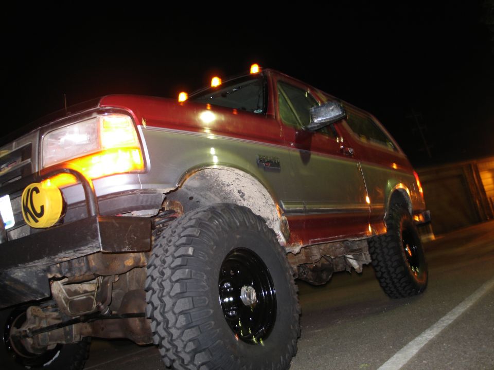 Red and silver Ford Bronco at night, low angle, mud tires, black wheels, KC light cover