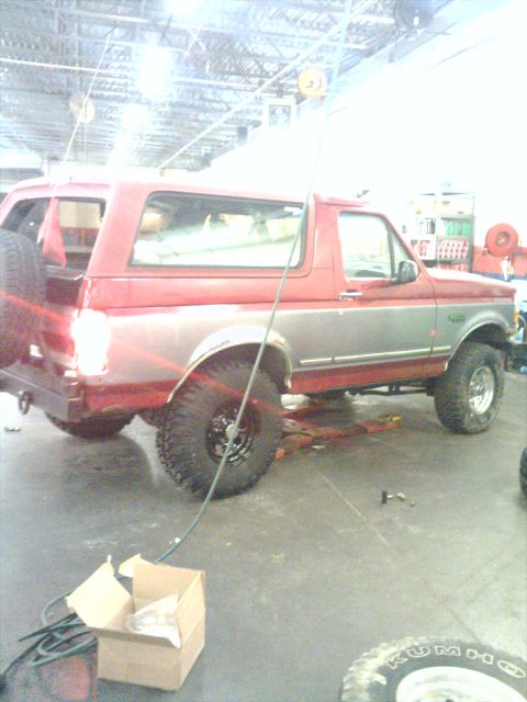 1995 Ford Bronco - Red-over-silver two-tone full-size Ford Bronco sitting inside a shop or warehouse bay, viewed from the driver's side rear three-quarter wit…