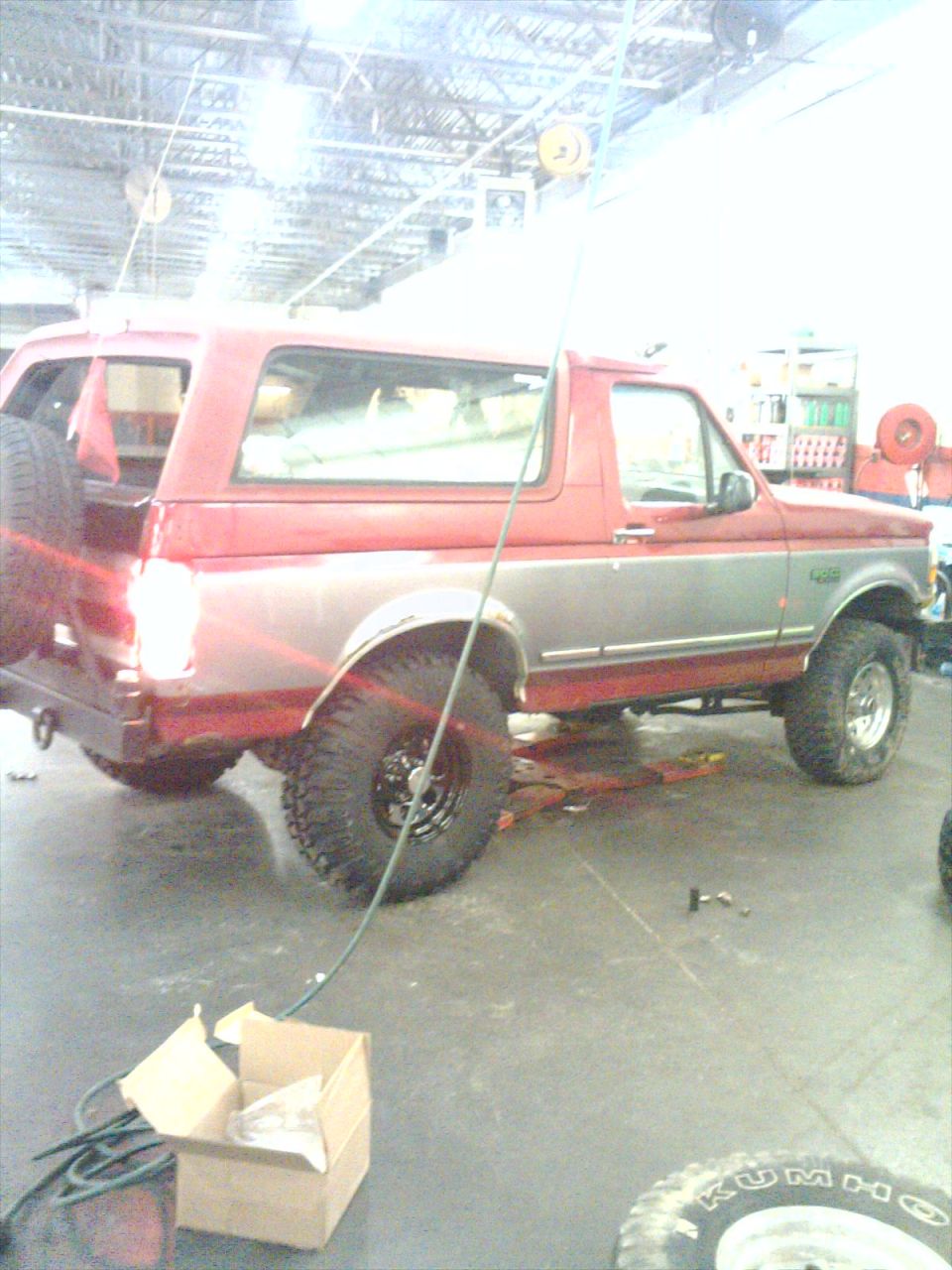 Red and silver Ford Bronco in a shop bay with a spare tire leaning against the rear fender