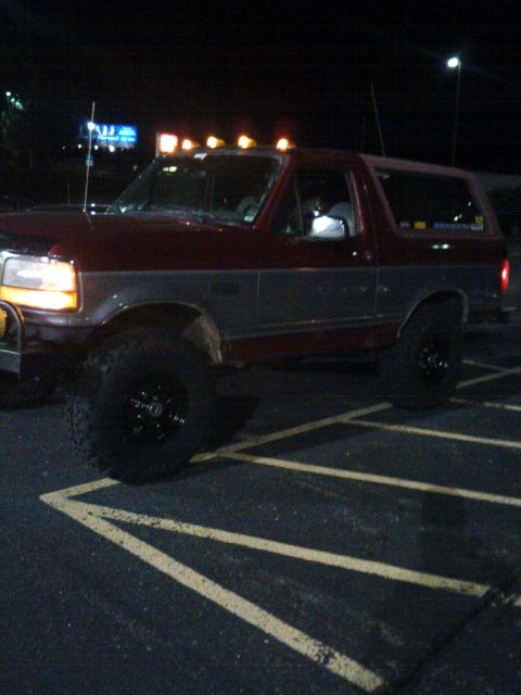 1995 Ford Bronco - Night shot of a lifted two-tone maroon-over-silver full-size Ford Bronco parked in a striped lot, headlights on and five amber roof cab lig…