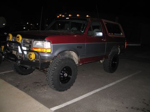 1995 Ford Bronco - Night flash shot of a lifted two-tone maroon-over-grey 1990s Ford Bronco angled in a parking stall, sitting on aggressive mud-terrain tires…