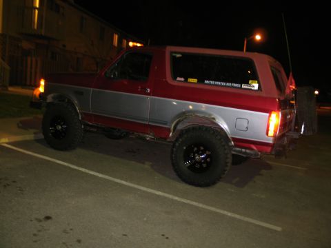 1995 Ford Bronco - A nighttime side view of a lifted two-tone red-over-silver full-size Ford Bronco in a parking lot, riding on black wheels with mud-terrain…